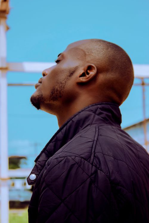 Free From below side view of brutal African American bald bearded male in stylish jacket standing with closed eyes under blue sky near metal construction Stock Photo