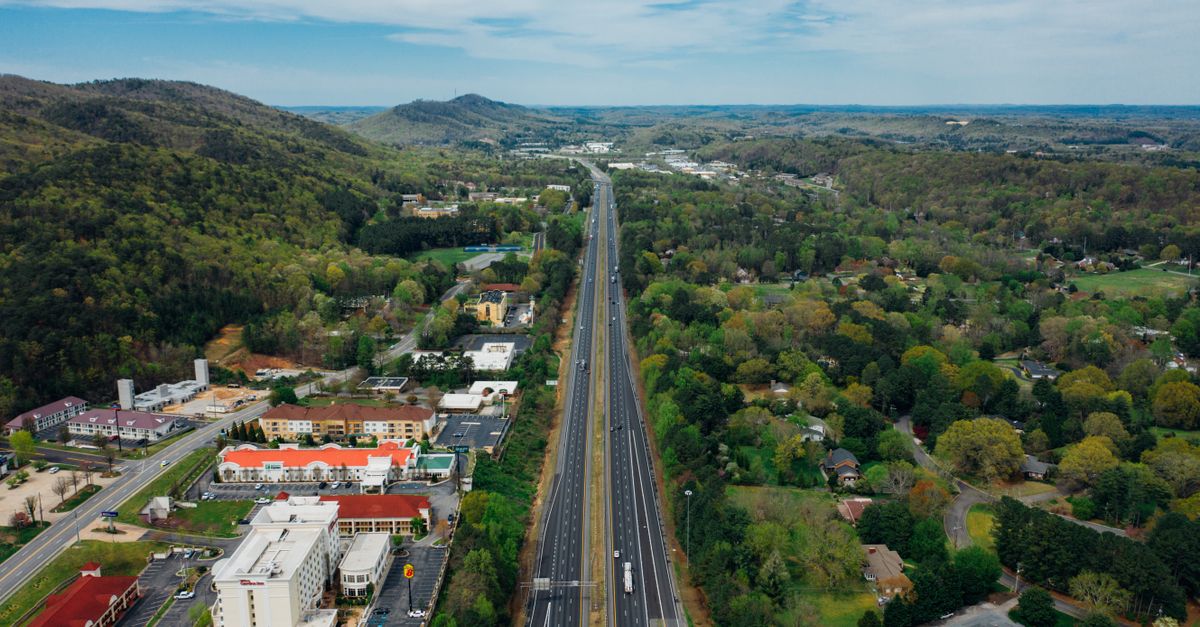 Drone view of narrow asphalt road with driving vehicles surrounded by green forest on hillside and colorful houses in city under spectacular endless sky