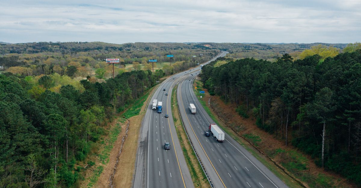 Aerial view of wavy asphalt roads with driving cars and trucks between bright trees under endless cloudy sky in rural zone