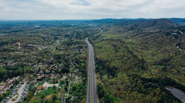 Drone view of narrow wavy asphalt road with driving cars surrounded by lush green trees and parked vehicles in town under sky with horizon line