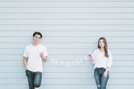 Content young Asian married couple in casual clothes holding garland with Mr and Mrs standing near blue wall and looking at camera