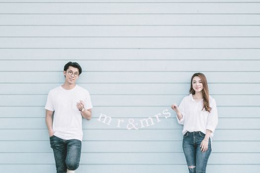 Content young Asian married couple in casual clothes holding garland with Mr and Mrs standing near blue wall and looking at camera