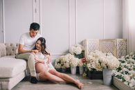 Happy young Asian male and pregnant female sitting together among white flowers in elegant living room
