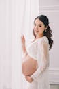 愉快的年轻的亚洲怀孕的女性在白色穿站立在窗帘附近和看照相机