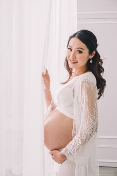 一位快乐的年轻亚洲孕妇穿着白色服装，站在窗帘旁看着镜头