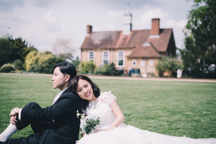 Content Newlywed Asian Couple Resting On Meadow Against Cozy Mansion