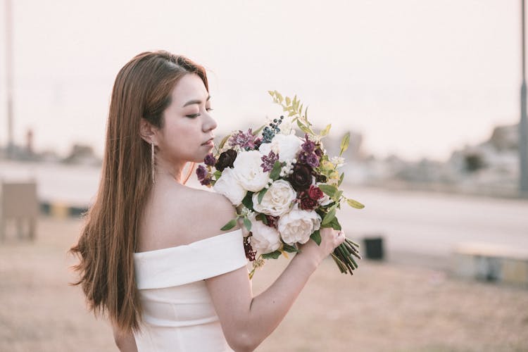 Young Calm Ethnic Bride Holding Wedding Bouquet In City Park