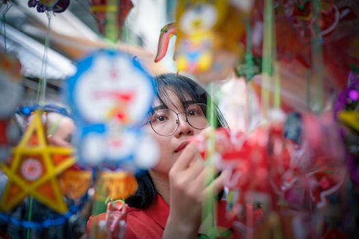 Asian woman with eyeglasses explores colorful hanging lanterns at an outdoor market.