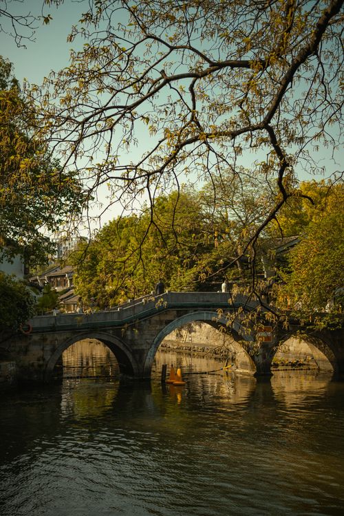 Free Historic stone bridge with arched design over a calm river, framed by lush trees. Stock Photo