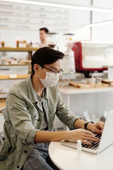 An adult man wearing a face mask works remotely on a laptop in a coffee shop.