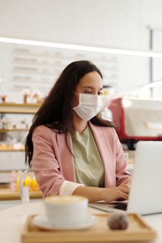 一位戴着口罩的成年女性正在一家现代咖啡馆里使用笔记本电脑