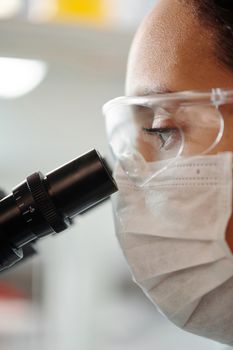 在实验室里，一位戴着面具和眼镜的科学家正在用显微镜仔细检查样品