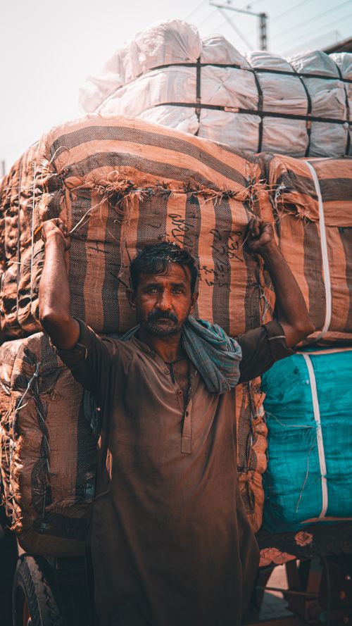 Free A hardworking laborer in traditional attire carrying a large bundle of goods outdoors in warm daylight. Stock Photo