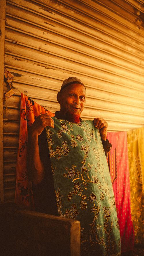 Free An elderly man showcasing colorful fabric at a market with warm sunlight Stock Photo