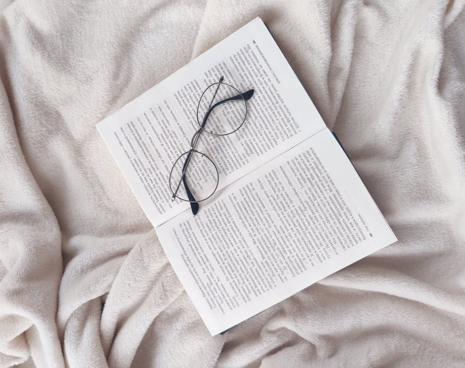 Free From above of opened book and eyeglasses placed on soft fleece white blanket in bedroom Stock Photo