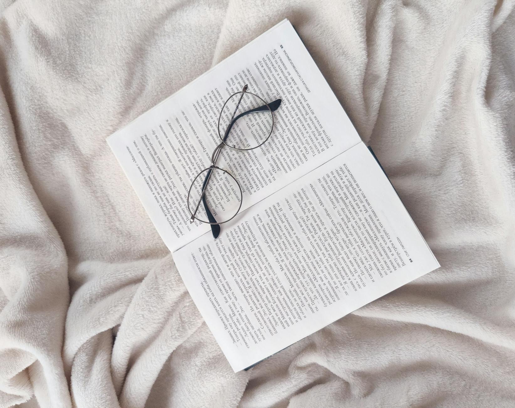 From above of opened book and eyeglasses placed on soft fleece white blanket in bedroom