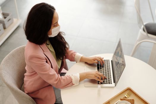 在现代室内环境中，一位戴着口罩的女士正用笔记本电脑工作，体现了新的正常工作习惯