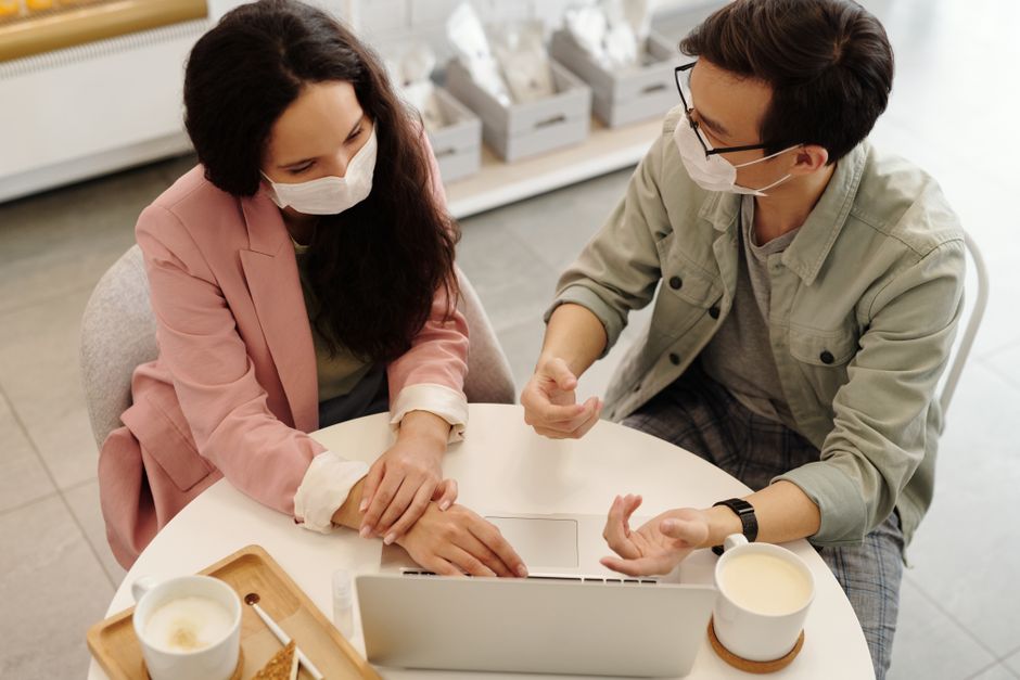 the importance of digital etiquette in hybrid workplaces - Two colleagues in masks discuss a project over a laptop, embodying modern work norms.