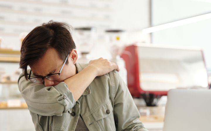 Adult man sneezing into his elbow inside a modern cafe while using a laptop.