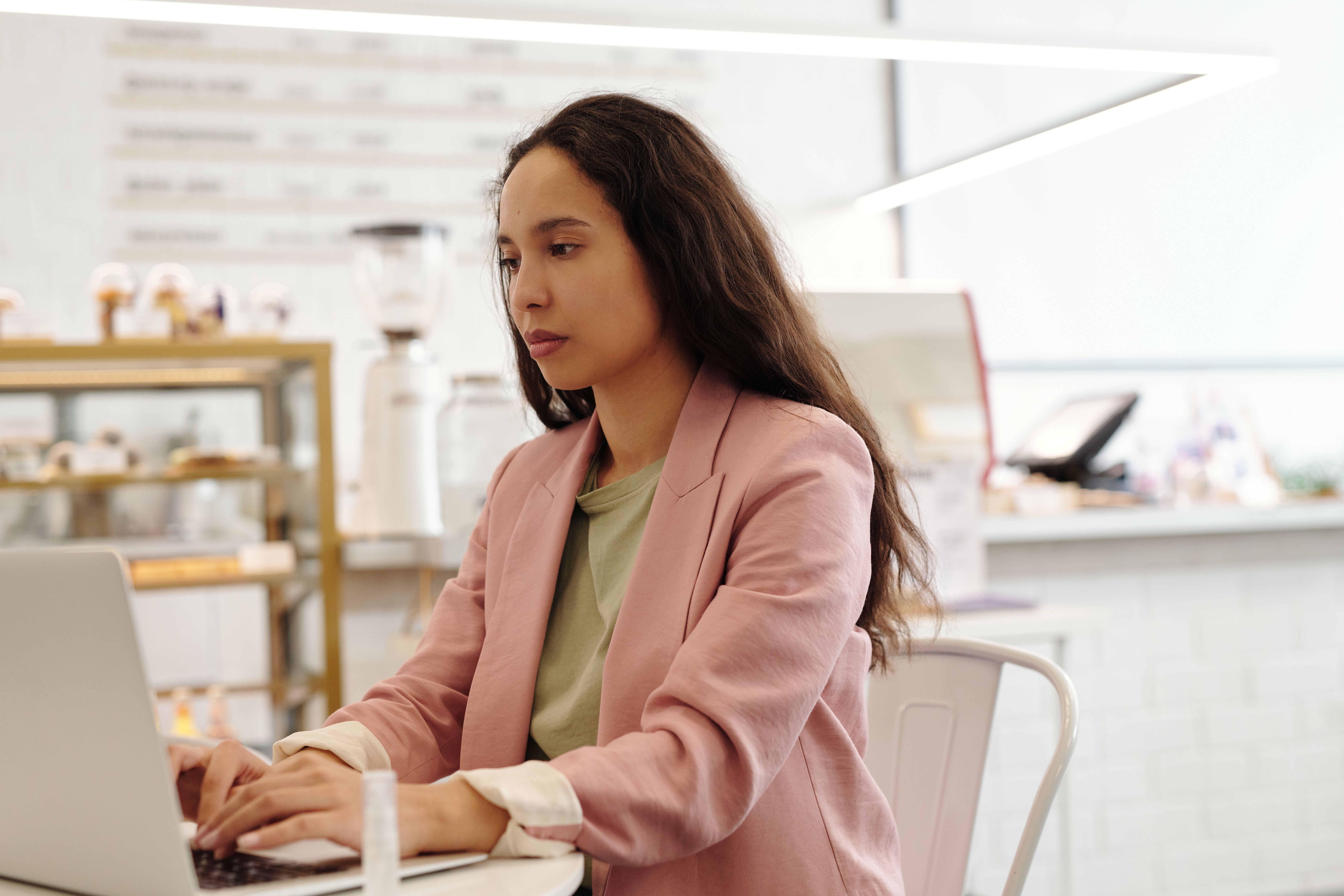 Mulher focada em um café trabalhando em um laptop, vestindo um blazer rosa.