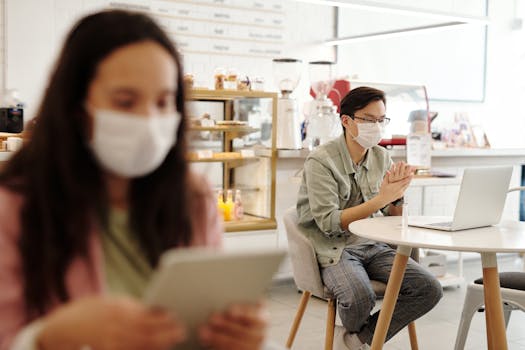 Adults wearing masks in a cafe, using devices, practicing social distancing.