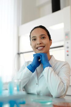 一位面带微笑的女科学家穿着白色实验服，坐在摆满试管的实验室里