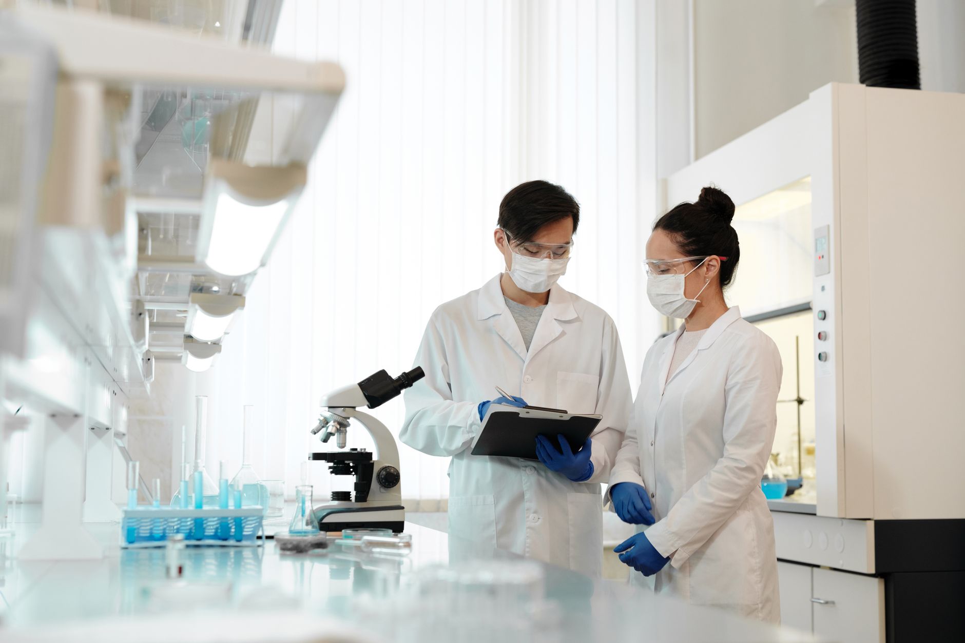 Two scientists in a laboratory wearing masks, discussing research with a microscope.