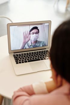 一名戴口罩的男子在室内挥手致意，他正用笔记本电脑进行视频通话