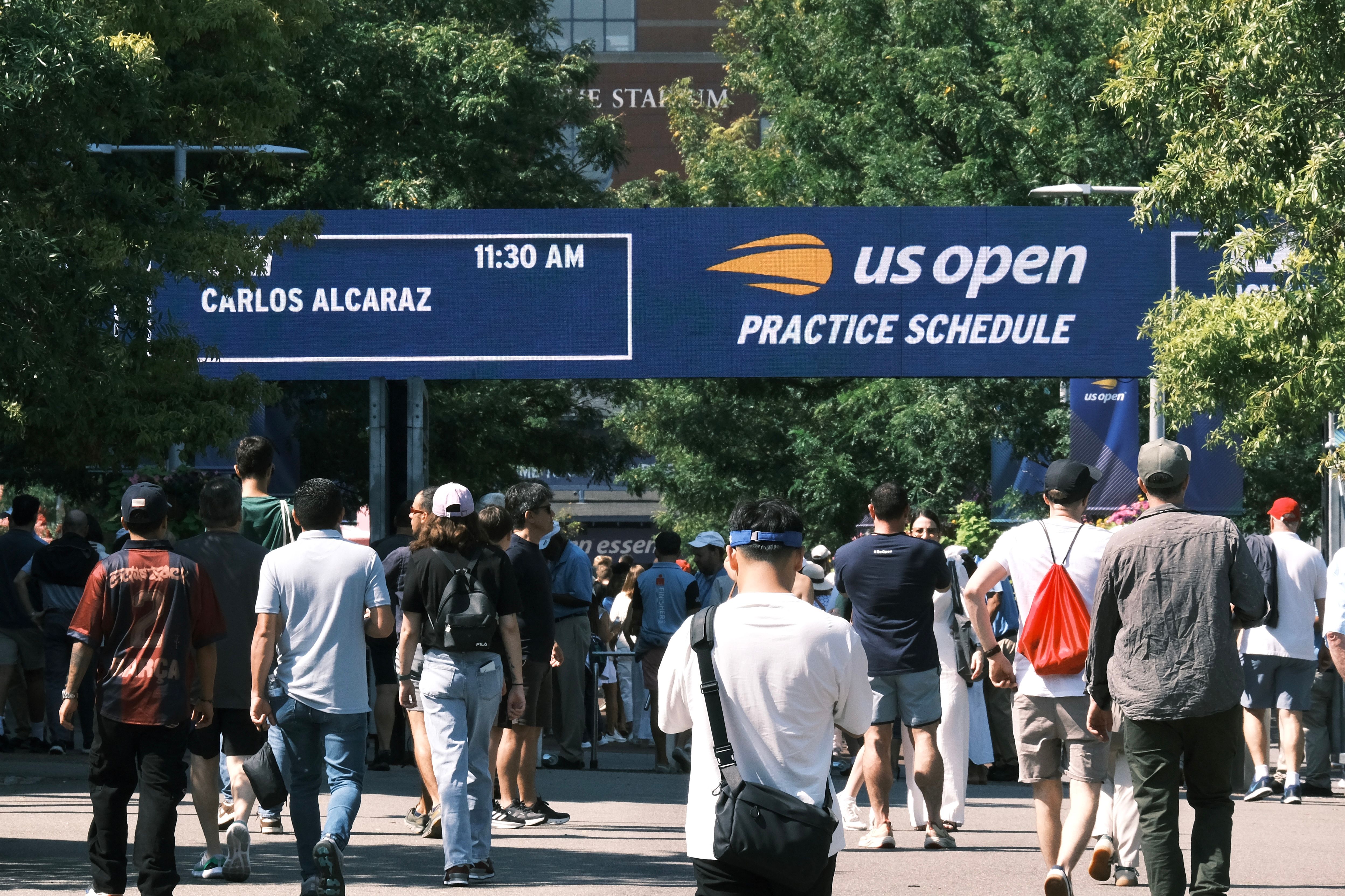 People gather at the US Open practice schedule board under clear skies.