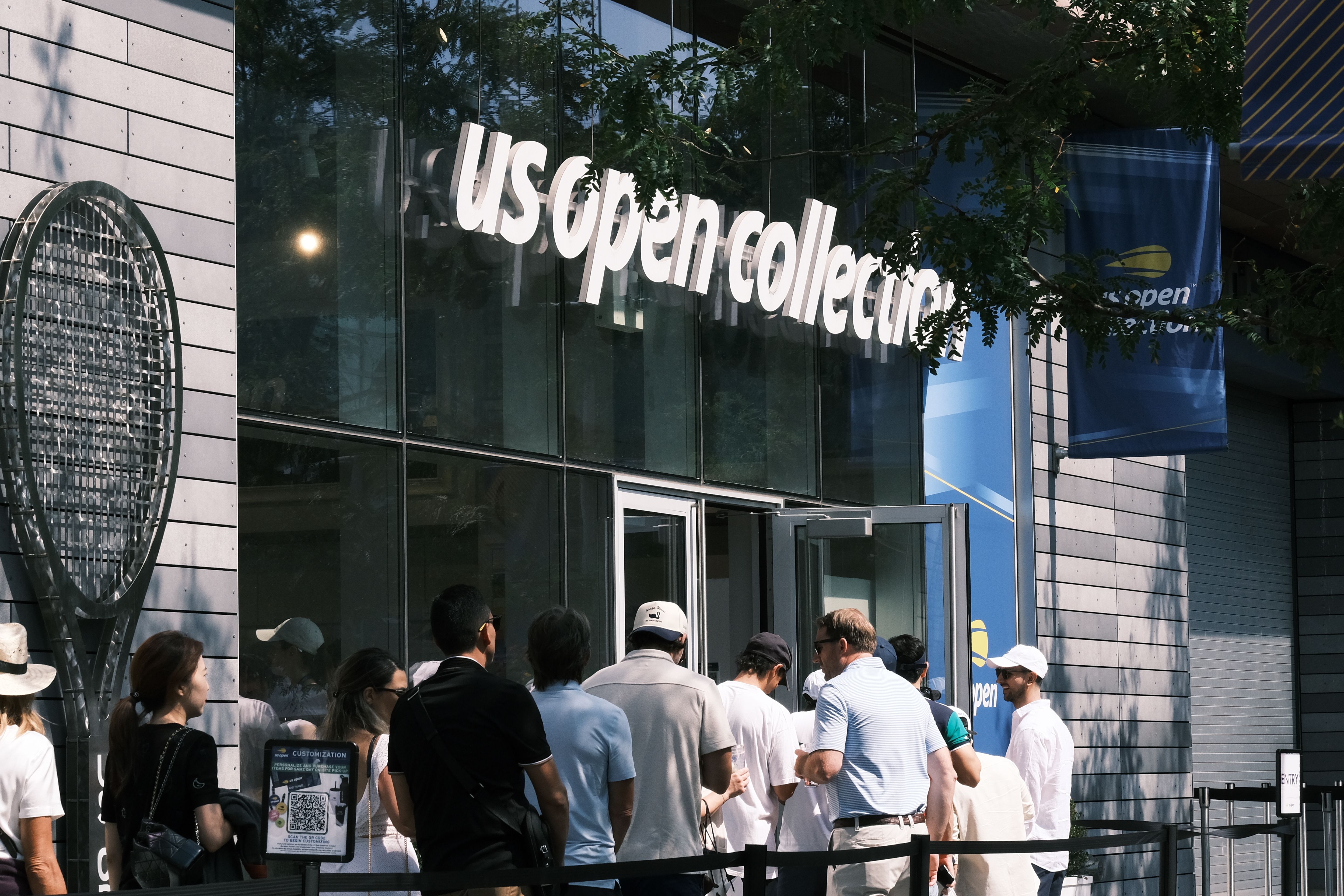Fotomural Gente entrando en la tienda US Open Collection, lo que muestra una escena comercial muy animada durante el evento de tenis.