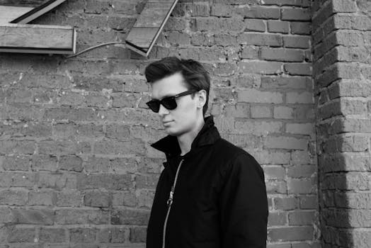 Stylish young adult in sunglasses leaning against a brick wall with a cool demeanor.