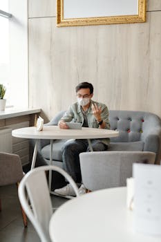 Young Asian man wearing a face mask while using a tablet for video calling indoors.