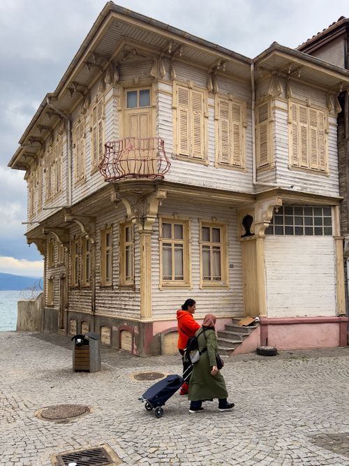 Free Two people walk past a traditional wooden house by the sea, showcasing unique architecture. Stock Photo