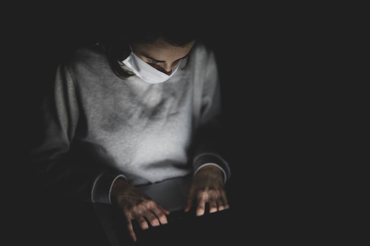 Man With Face Mask Using Laptop In The Dark