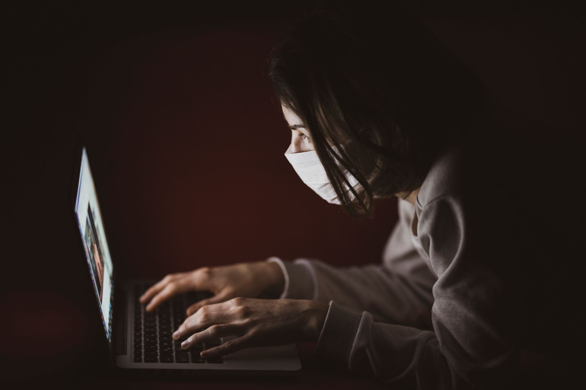 ai news today - A woman wearing a face mask focuses on her laptop screen in dim lighting, highlighting remote work.