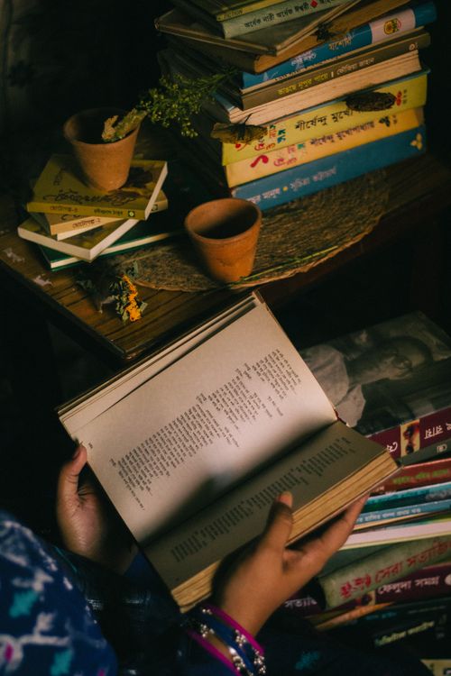 Free Hands holding a Bengali book in a cozy room with stacks of books and clay pots. Stock Photo