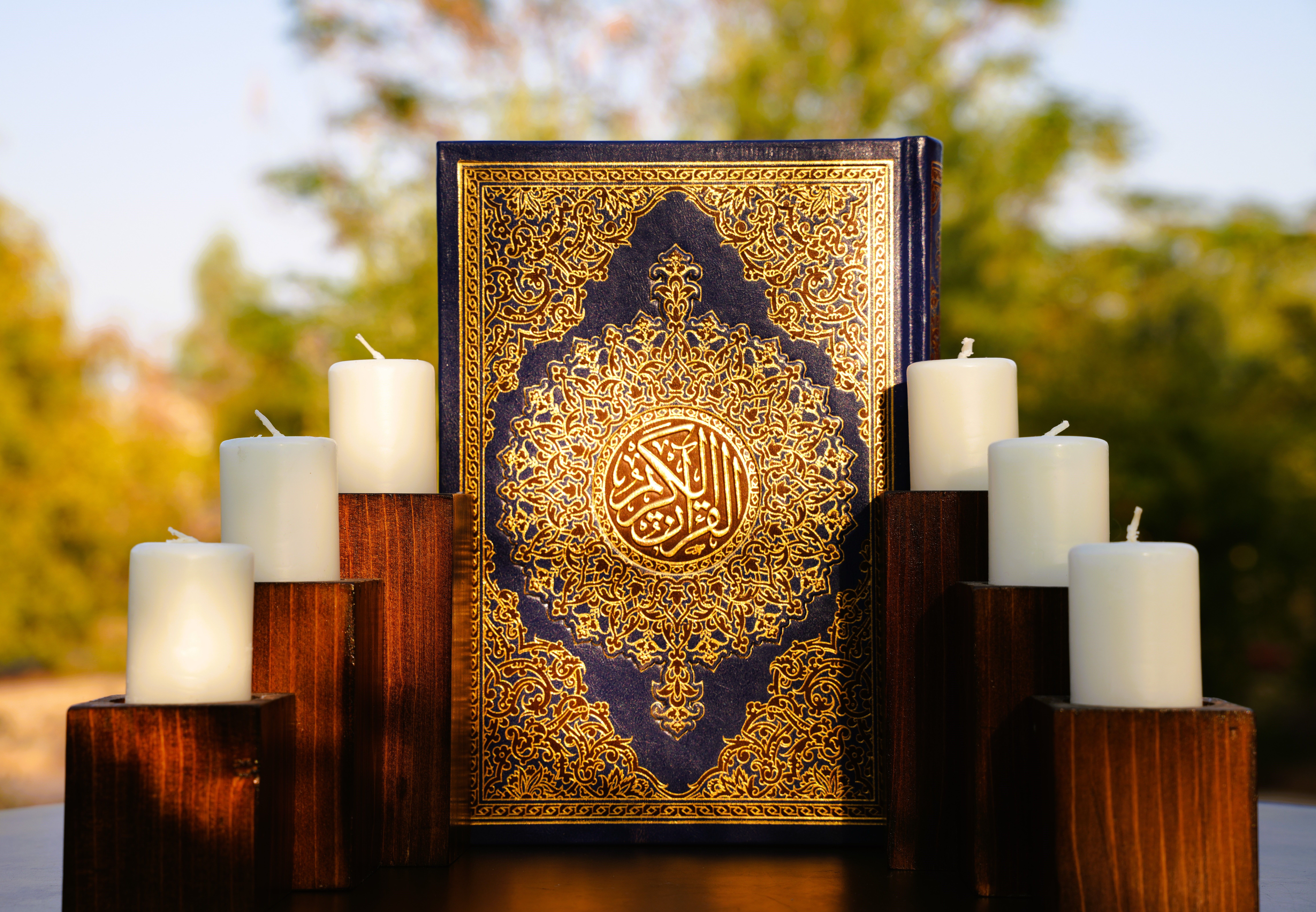 A beautifully ornate Quran surrounded by white candles, set outdoors in the sunlight, symbolizing peace and spirituality.