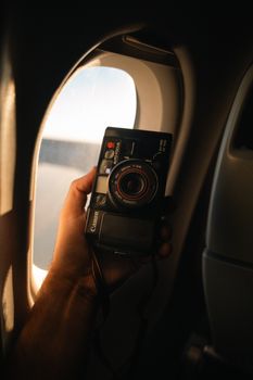 A hand holding a vintage camera by an airplane window with soft light.