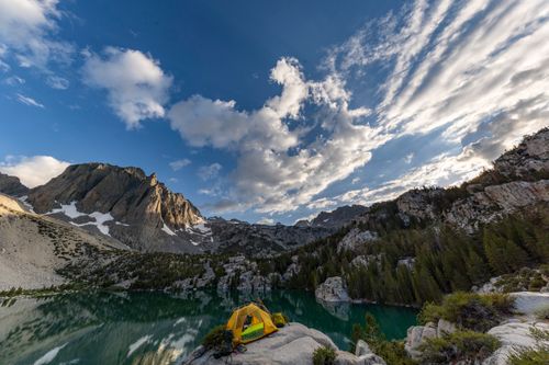 Free Outdoor camping scene by a pristine lake with a vibrant yellow tent and dramatic mountain view under a blue sky. Stock Photo