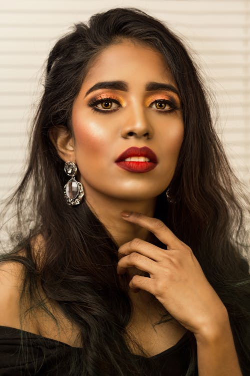 Portrait of gorgeous young ethnic lady with long dark hair and makeup touching neck and looking at camera