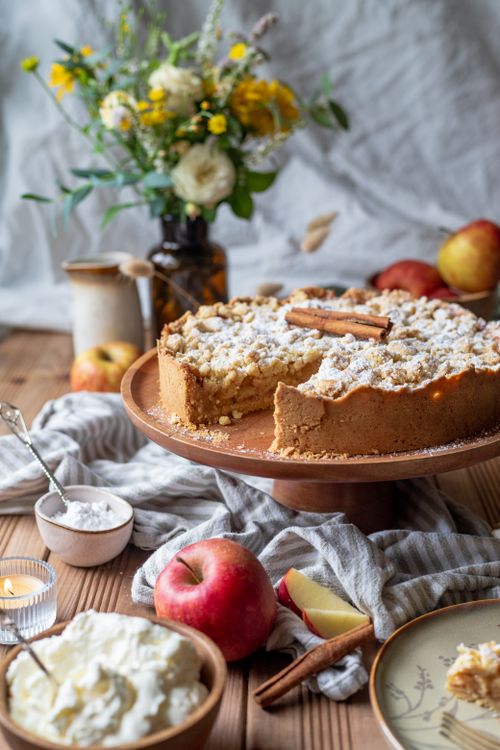 Free A freshly baked apple crumble tart on a wooden stand, surrounded by ingredients and flowers, creates a cozy, inviting scene. Stock Photo