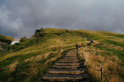 Free Hikers ascending a rustic path towards Skógafoss waterfall amidst lush Icelandic scenery. Stock Photo