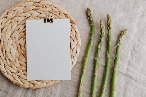 From above of four fresh green asparagus sprouts and blank sheet of paper over round wicker placemat laid on white tablecloth