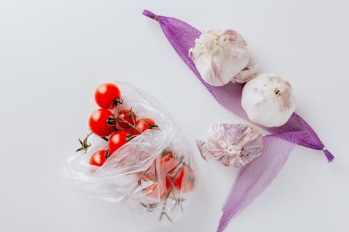 Top view of fresh cherry tomatoes in transparent polyethylene bag and three heads of garlic placed on purple grid isolated on white background