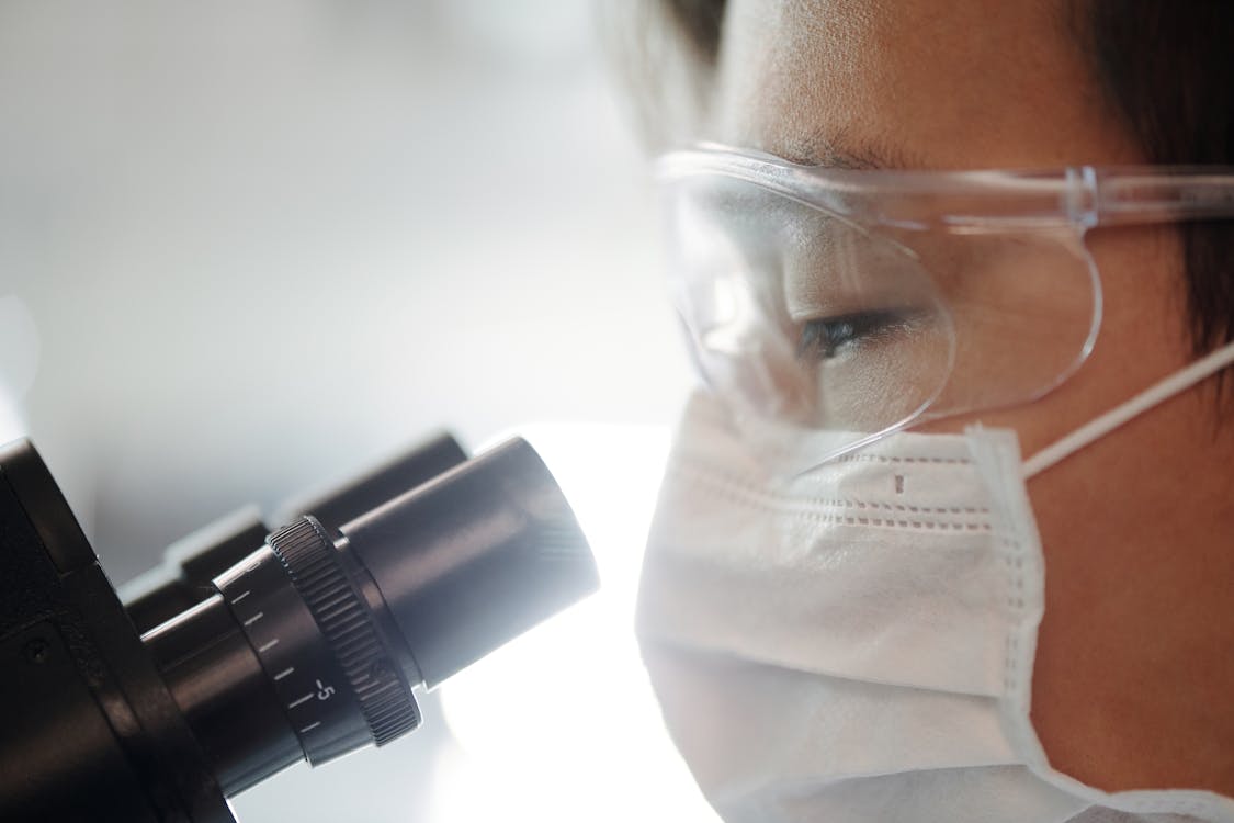 Man wearing goggles and a mask looks through a microscope.