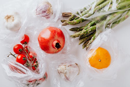 Top view of pomegranate in center surrounded by bundle of raw asparagus with orange and bunch of tomatoes put near heads of garlic in plastic bags on white surface