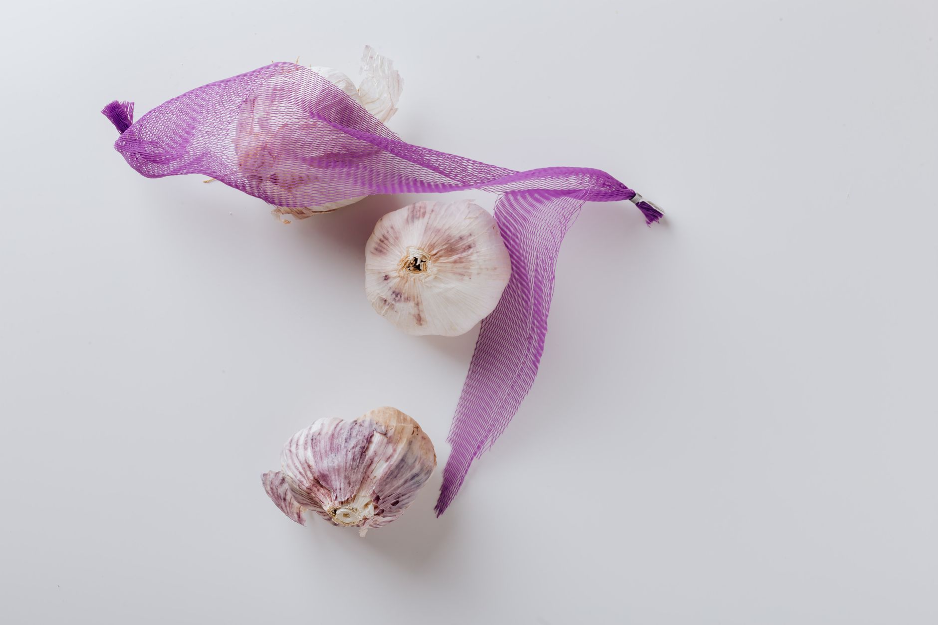 Top view of whole heads of garlic with dry clean husk partially covered with violet net fixed with metal pieces on edges and put on white surface