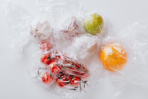 Free From above of bunch of ripe tomatoes put near garlic bulbs and citrus fruits covered with transparent plastic bags on white surface Stock Photo