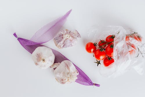 From above of bunch of ripe tomatoes put in plastic bag near raw heads of garlic put on violet transparent net on white surface