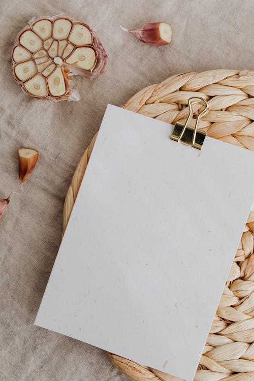 Top view of sheet of white blank paper with metal clip composed on straw table mat among cut cloves of garlic